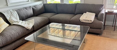 A living room with a sectional sofa, a glass coffee table, and a view through large windows.