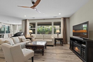 Ein Wohnzimmer mit Deckenventilator, Kamin, Fernseher und einem großen Fenster mit Vorhängen.
