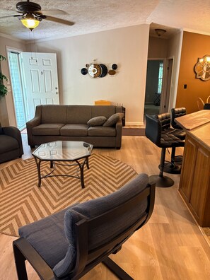 A living room with a sofa, a coffee table, a chair, a dining area with a bar, and a ceiling fan. - 3-bedroom, 6-beds house in Asheville close to downtown. Sleeps 8. Peaceful... (Asheville)