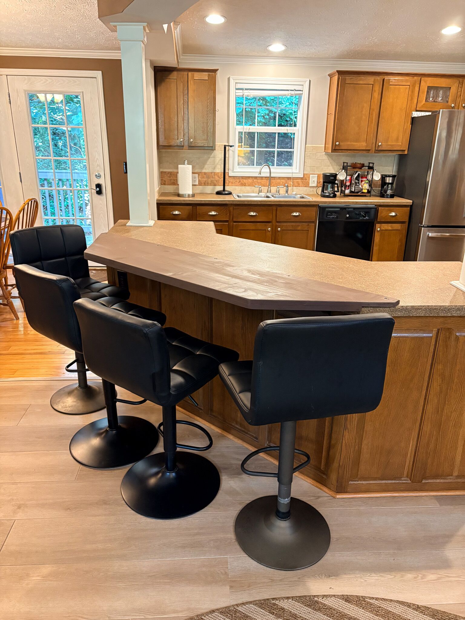 A kitchen with a large island, wooden cabinets, a stainless steel refrigerator, and black bar stools.