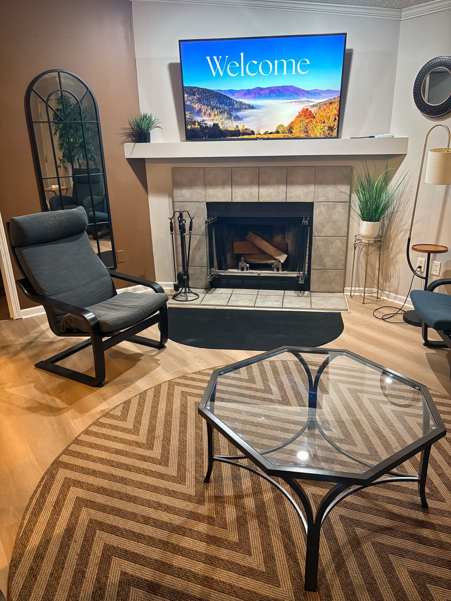 A modern living room with a fireplace, a television displaying 'Welcome,' and a glass coffee table.