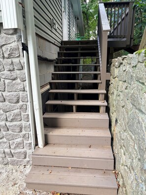 A set of wooden exterior stairs leading to a house with a stone wall on one side and a metal railing on the other.