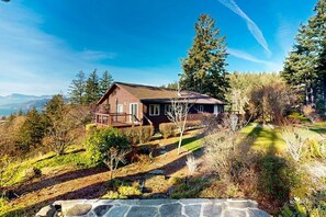 A two-story house with a stone foundation, surrounded by a well-maintained garden and tall trees. - Gorge Retreat | Panoramic Views & Private Trail (Stevenson)