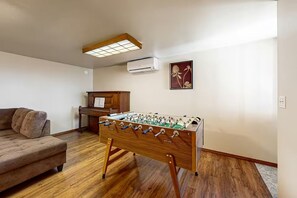 A room with a wooden foosball table, a sofa, a wooden cabinet, and a wall-mounted air conditioner. - Gorge Retreat | Panoramic Views & Private Trail (Stevenson)