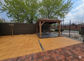 Un área de estar al aire libre con techo, suelo de ladrillo, pérgola de madera y cerca de metal.