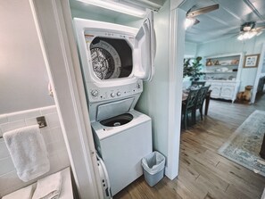 A compact laundry room with a front-loading washer and dryer, a small trash bin, and a towel rack.