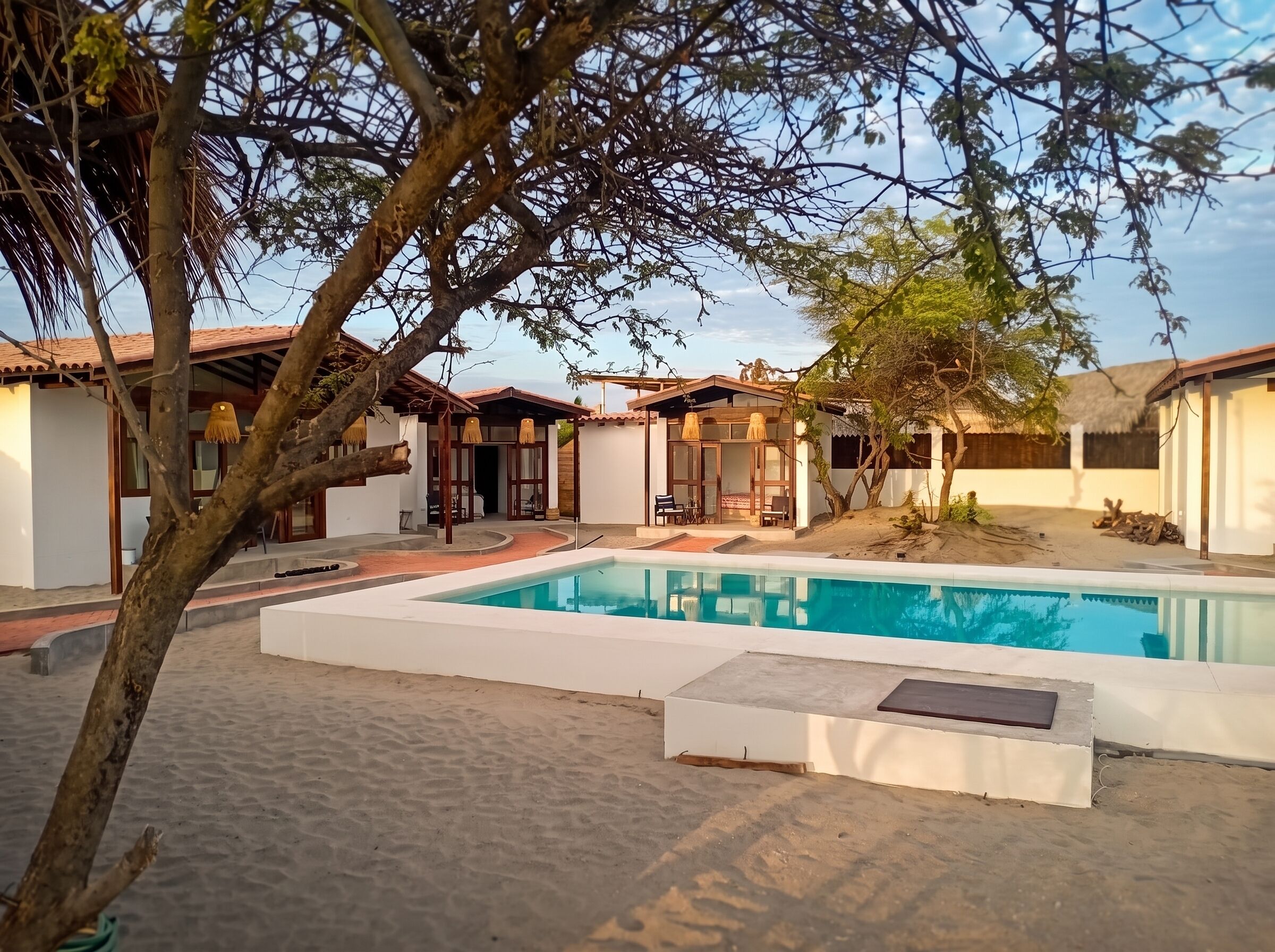 A pool area with a clear blue pool, surrounded by a sandy beach and a few trees.