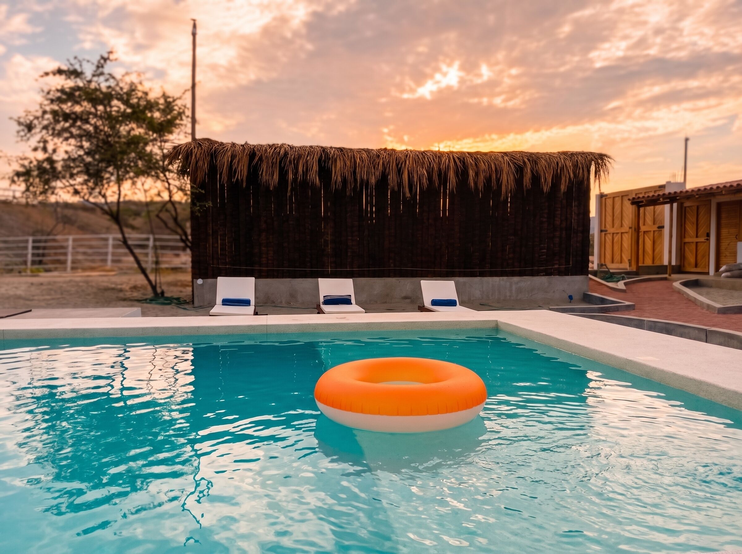 A swimming pool with a clear blue water surface, a floating orange ring, and a thatched roof structure in the background.