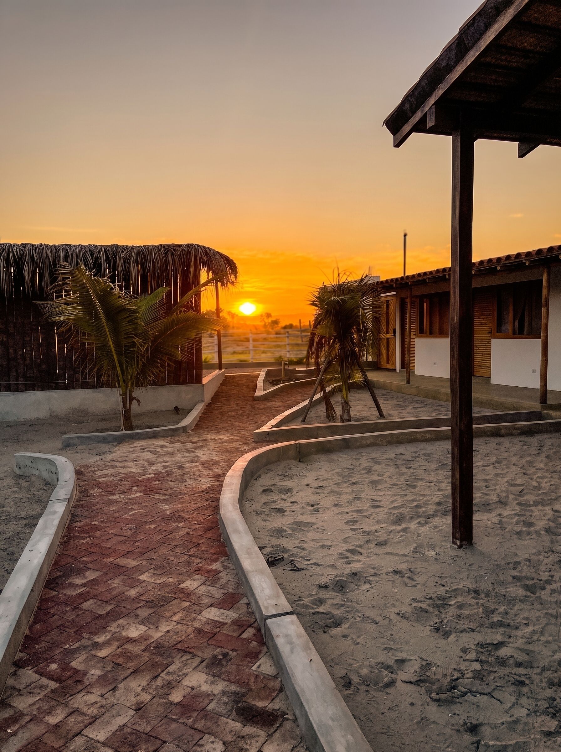 A pathway leading to a building with a thatched roof and palm trees, surrounded by sand.
