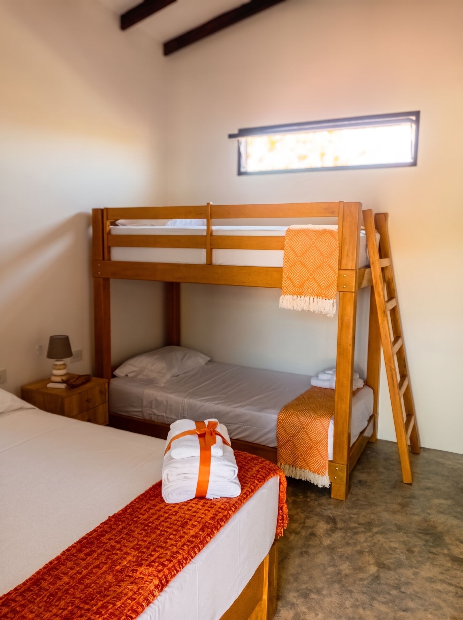 A bunk bed room with a window, a bedside table with a lamp, and a red throw blanket.