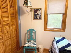 Een kamer met een houten garderobe, een blauwgroene stoel, een wandlamp, een ingelijst kunstwerk, een raam met zonwering, en een bed met een gestreept deken.
