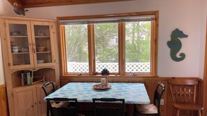 A dining area with a table set for four, wooden chairs, a cabinet with glass doors, and a window with a view of trees and a white fence. - Sweet split level 4 bedroom house, quiet neighborhood, walking distance to beach (Kitty Hawk)