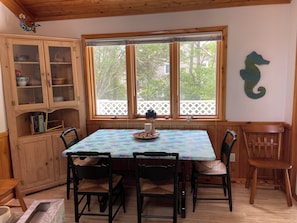 A dining area with a table set for four, wooden chairs, a cabinet with glass doors, and a window with a view of trees and a white fence. - Sweet split level 4 bedroom house, quiet neighborhood, walking distance to beach (Kitty Hawk)