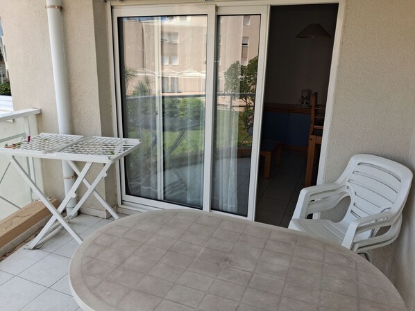 A balcony with a table and chairs, a sliding glass door, and a view of a building and greenery.