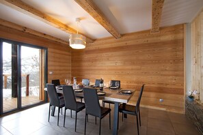 A dining area with a wooden interior, a large table set for six, and a view through glass doors. - Chalet rénové à 150 m du télésiège | Vue montagne (Mont-de-Lans)
