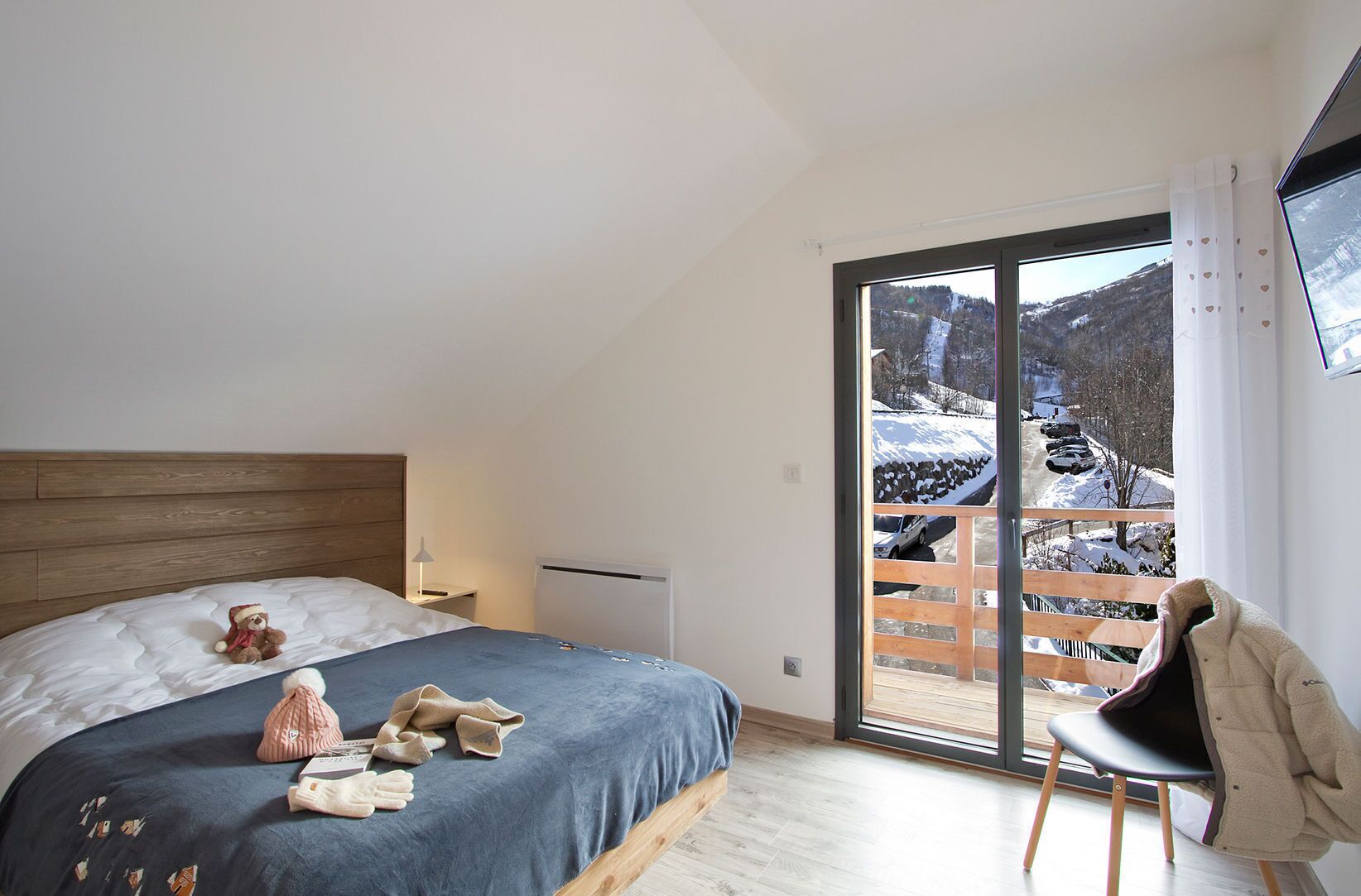 Een slaapkamer met een bed, een stoel, een teddybeer en uitzicht op een besneeuwd landschap door een groot raam.