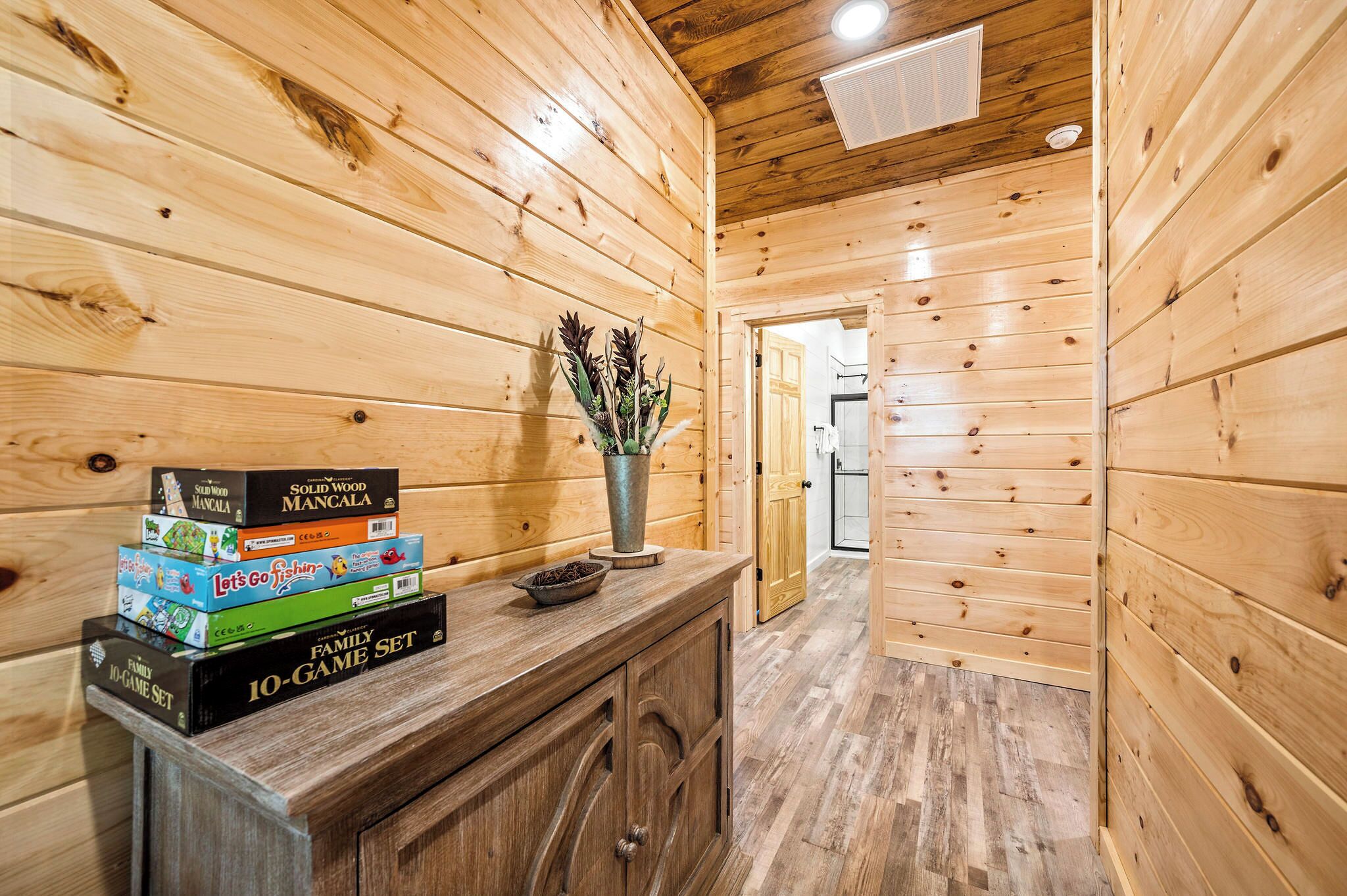 A wooden interior with a wooden cabinet, a vase with dried plants, and a hallway leading to another room.