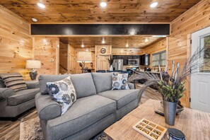 Ein gemütliches Wohnzimmer mit einem grauen Sofa, gemusterten Kissen und einem Couchtisch mit Fernbedienung und Deko-Objekten.