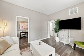 A modern living room with a white sofa, a coffee table, a flat-screen TV mounted on the wall, and a desk with a chair. - Charming One Bed with Office near Chicago! (Forest Park)