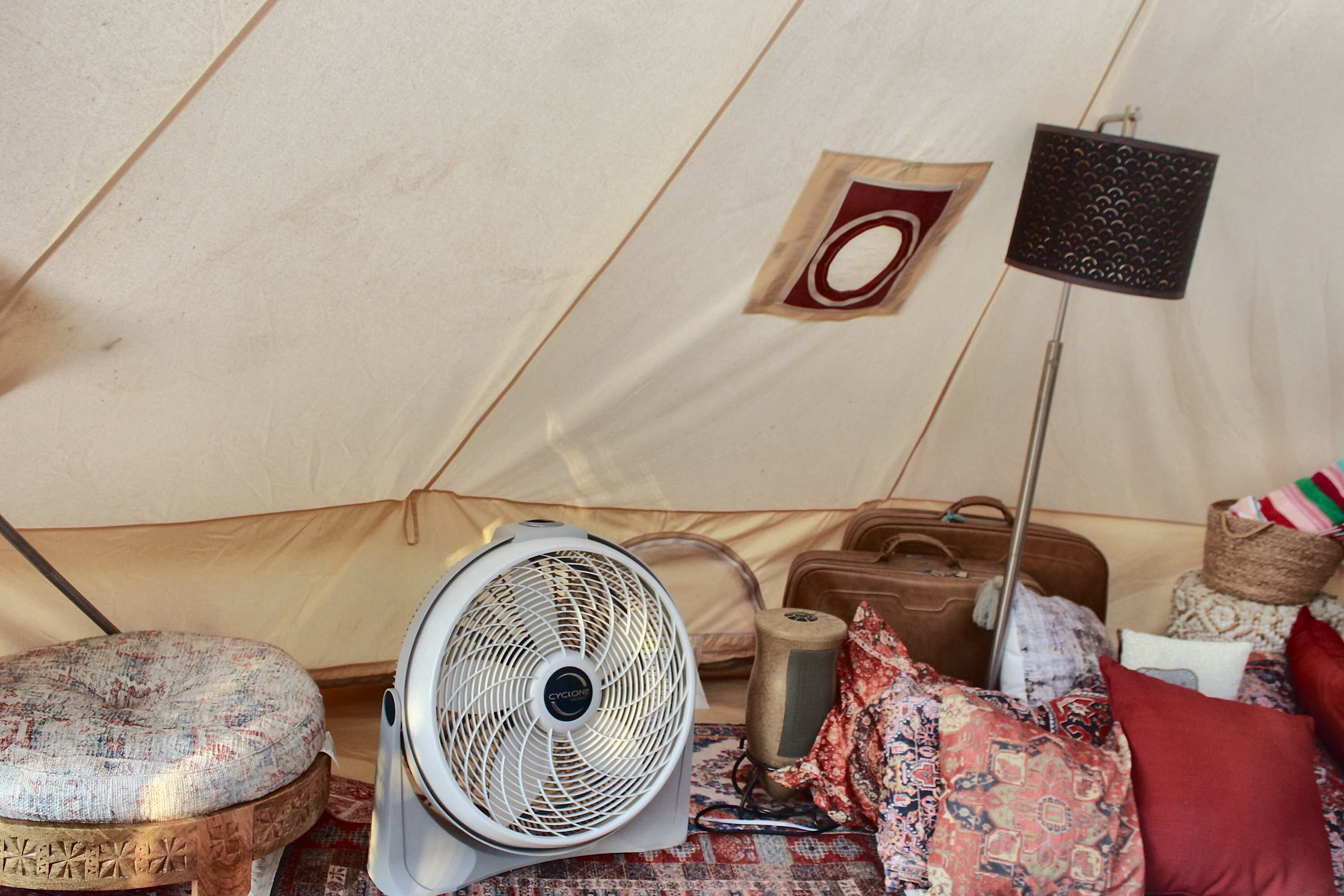 A tent interior with a standing fan, a small stool, a suitcase, and a floor lamp.
