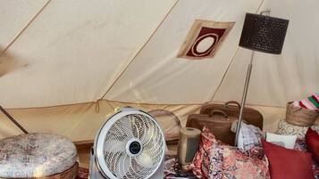 A tent interior with a standing fan, a small stool, a suitcase, and a floor lamp.