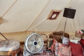 A tent interior with a standing fan, a small stool, a suitcase, and a floor lamp.