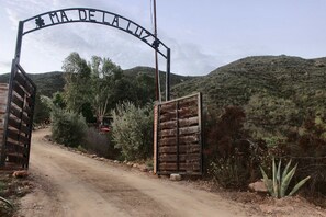 A rustic gate with a wooden structure and metal framework, leading to a dirt road surrounded by vegetation and hills.