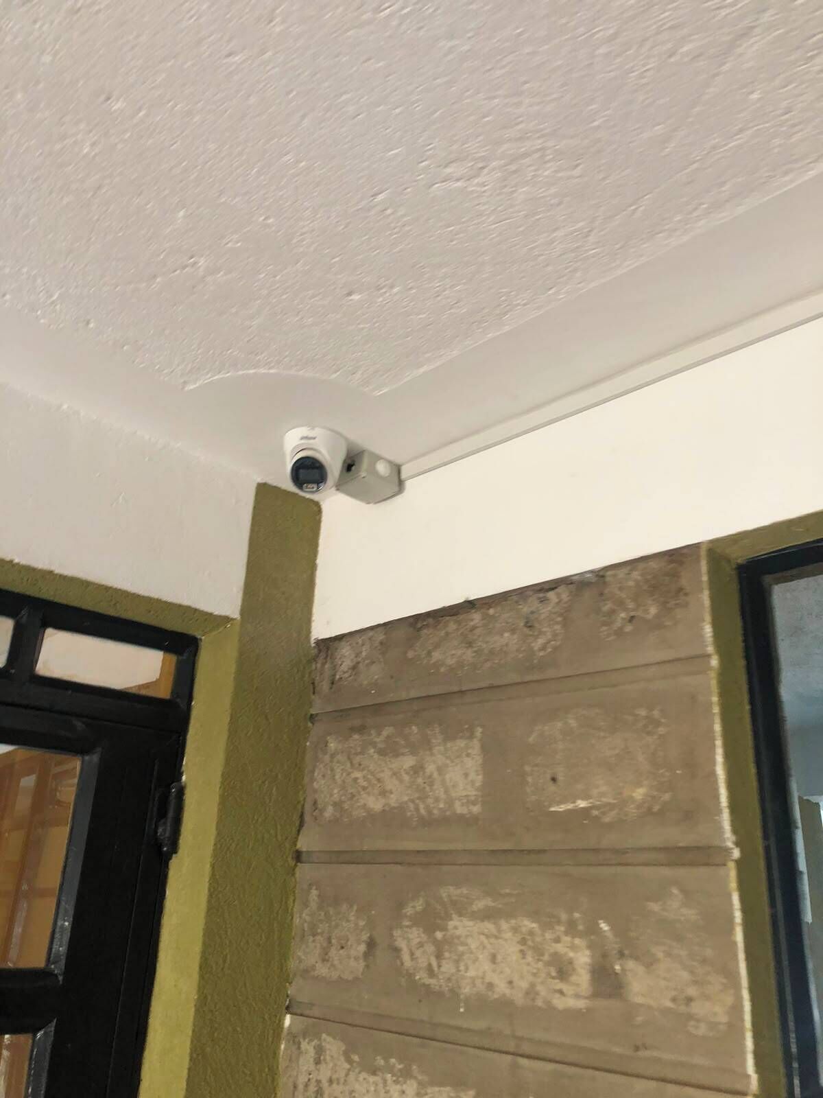 Una cámara de seguridad en un techo blanco, una puerta con marco negro y una pared con textura.