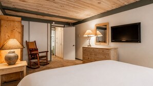 Ein Schlafzimmer mit einem Bett, einer Kommode, einem Sessel, einer Lampe, einem Fernseher und einer Tür zu einem anderen Raum.