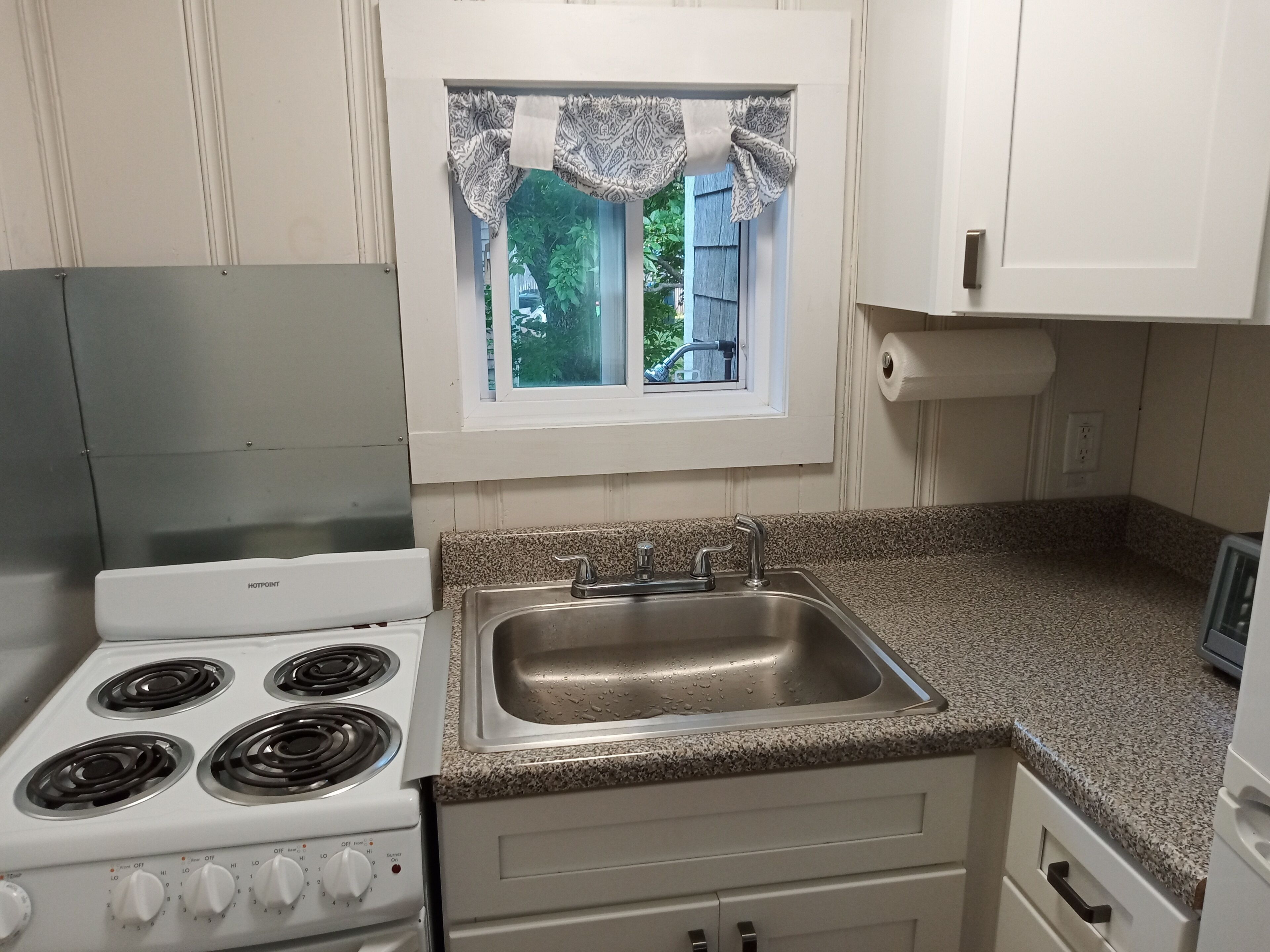 Una cocina con un horno blanco, un lavamanos de acero inoxidable y una ventana con un cortinero estampado.