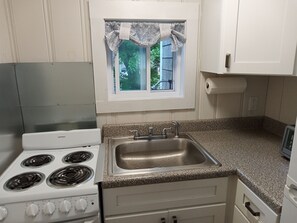 A kitchen with a white stove, a stainless steel sink, and a window with a patterned valance. - Cozy 2BR cottage, walk to beaches, restaurants, mini golf. (Yarmouth)