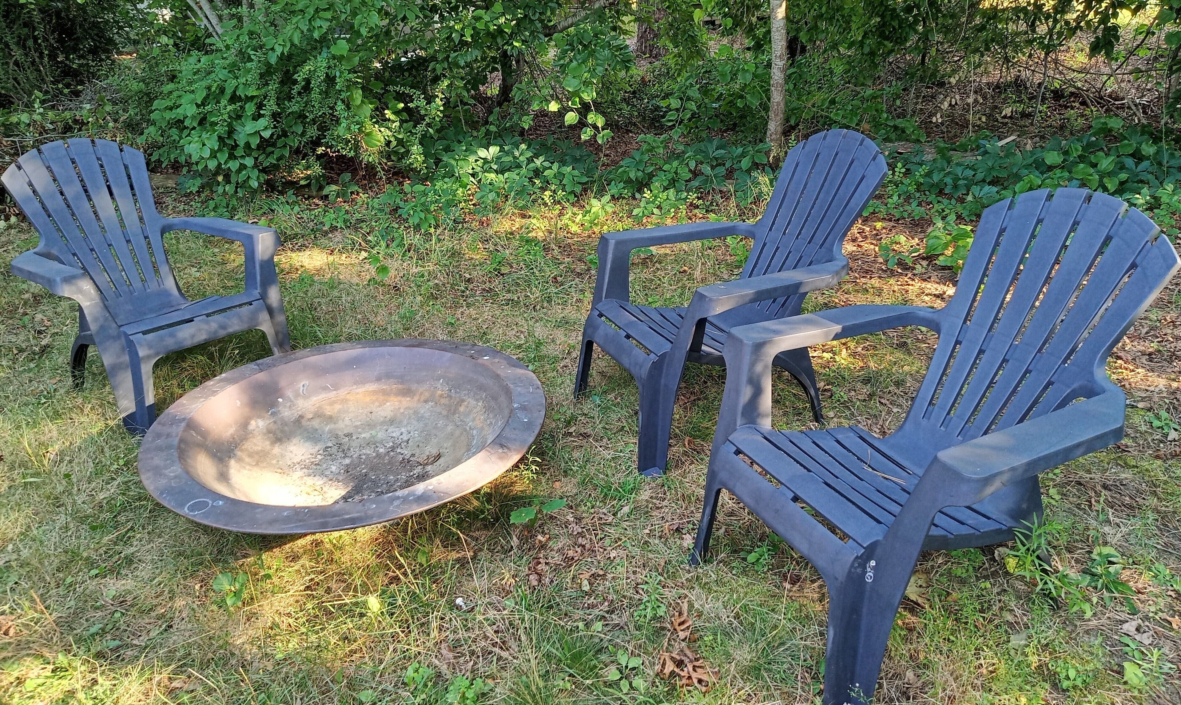 Tres sillas Adirondack y un brasero en un espacio al aire libre con césped, y vegetación densa al fondo.
