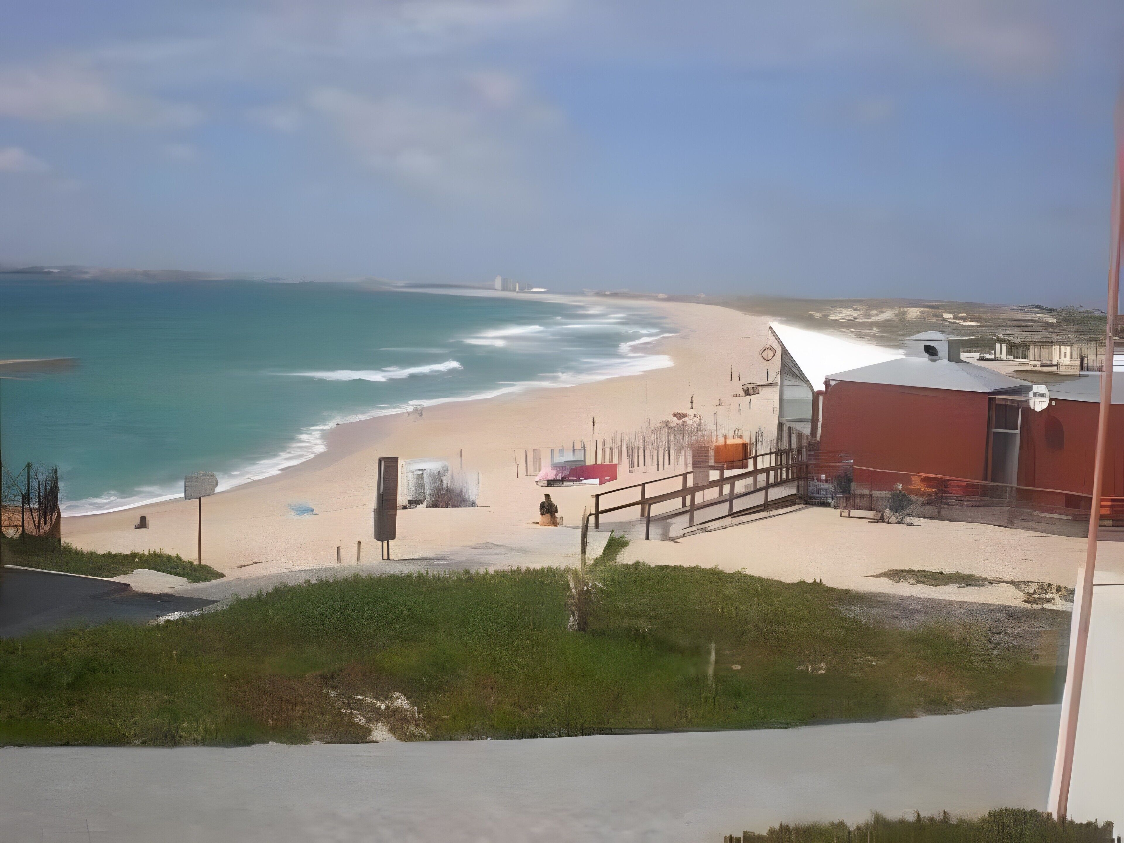 Vista da praia com um edifício, céu claro e mar calmo.