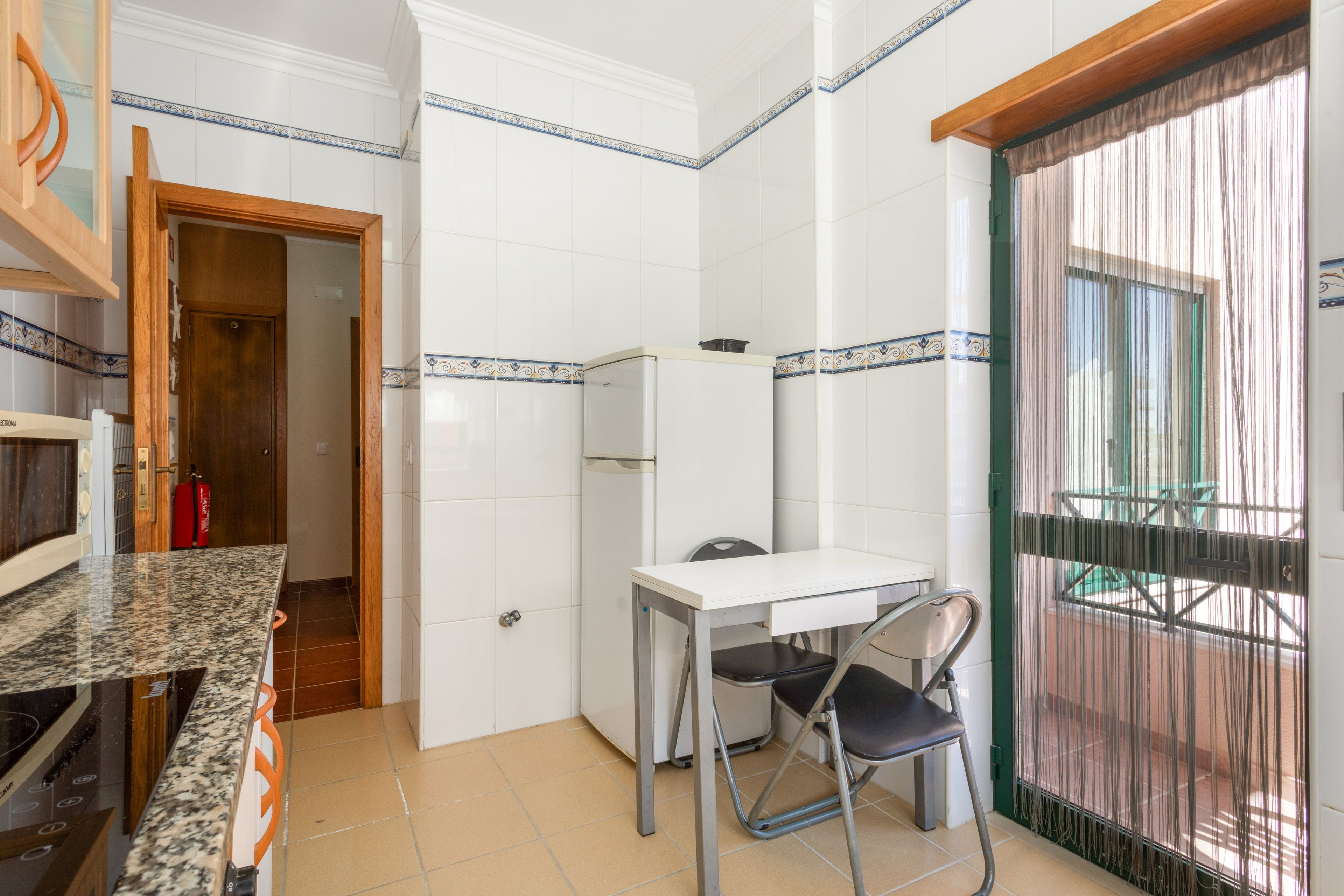 Cozinha compacta com azulejos brancos, bancada de granito, uma pequena mesa com uma cadeira e uma porta que dá para uma varanda.