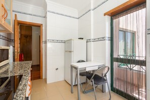 A compact kitchen with white tiles, a granite countertop, a small table with a chair, and a door leading to a balcony.