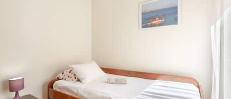 Un lit simple avec un cadre en bois, une couvre-lit blanche et un oreiller à motifs floraux. Il y a une petite table de chevet avec une lampe et, au mur, un tableau encadré représentant un bateau.