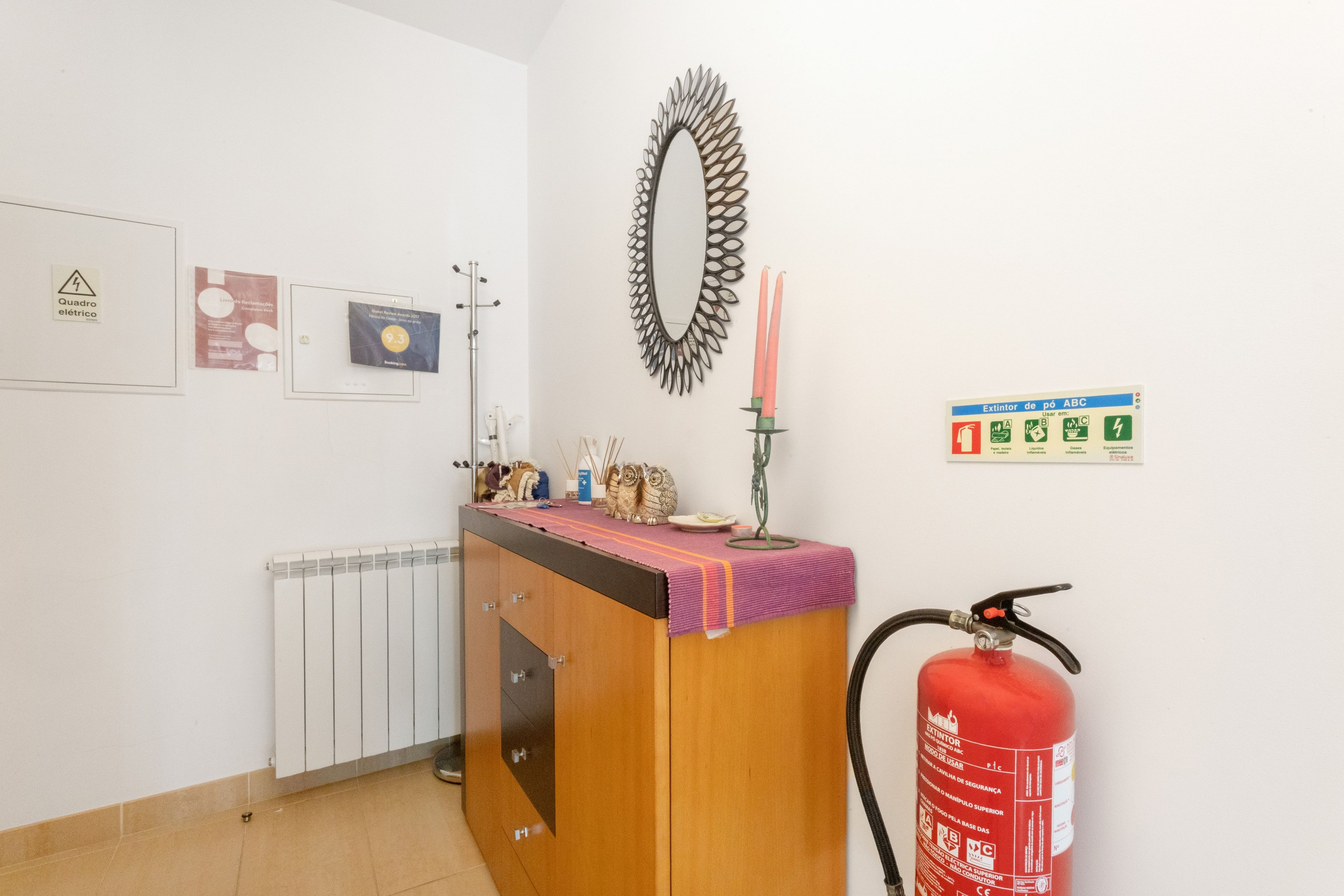 A reception area with a wooden counter, a fire extinguisher, a radiator, and various wall-mounted items including a mirror, framed certificates, and a safety sign.