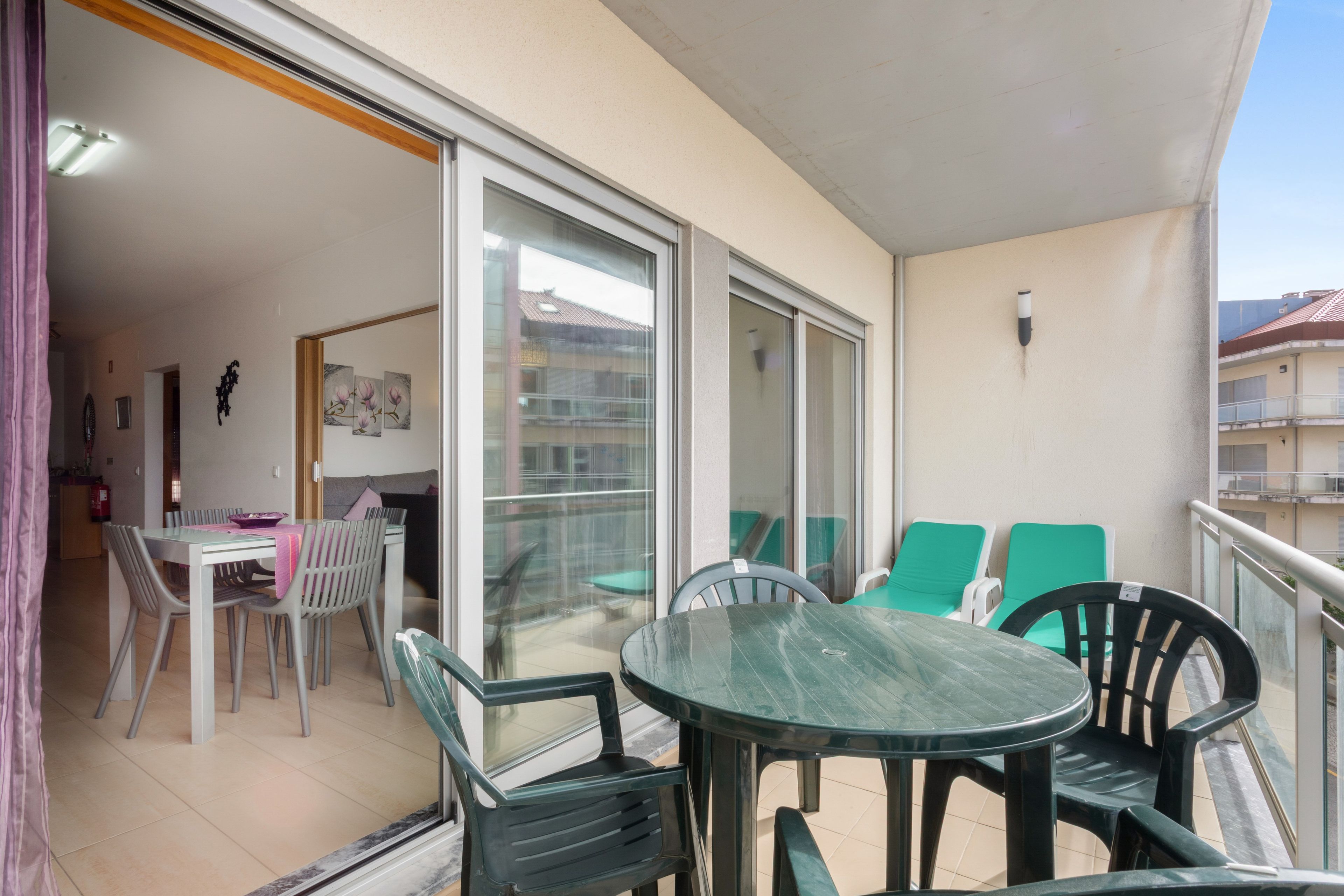 Un balcón al aire libre con una mesa y sillas, una puerta corredera de vidrio que da a un área de comedor interior, y vista de un edificio en la calle.