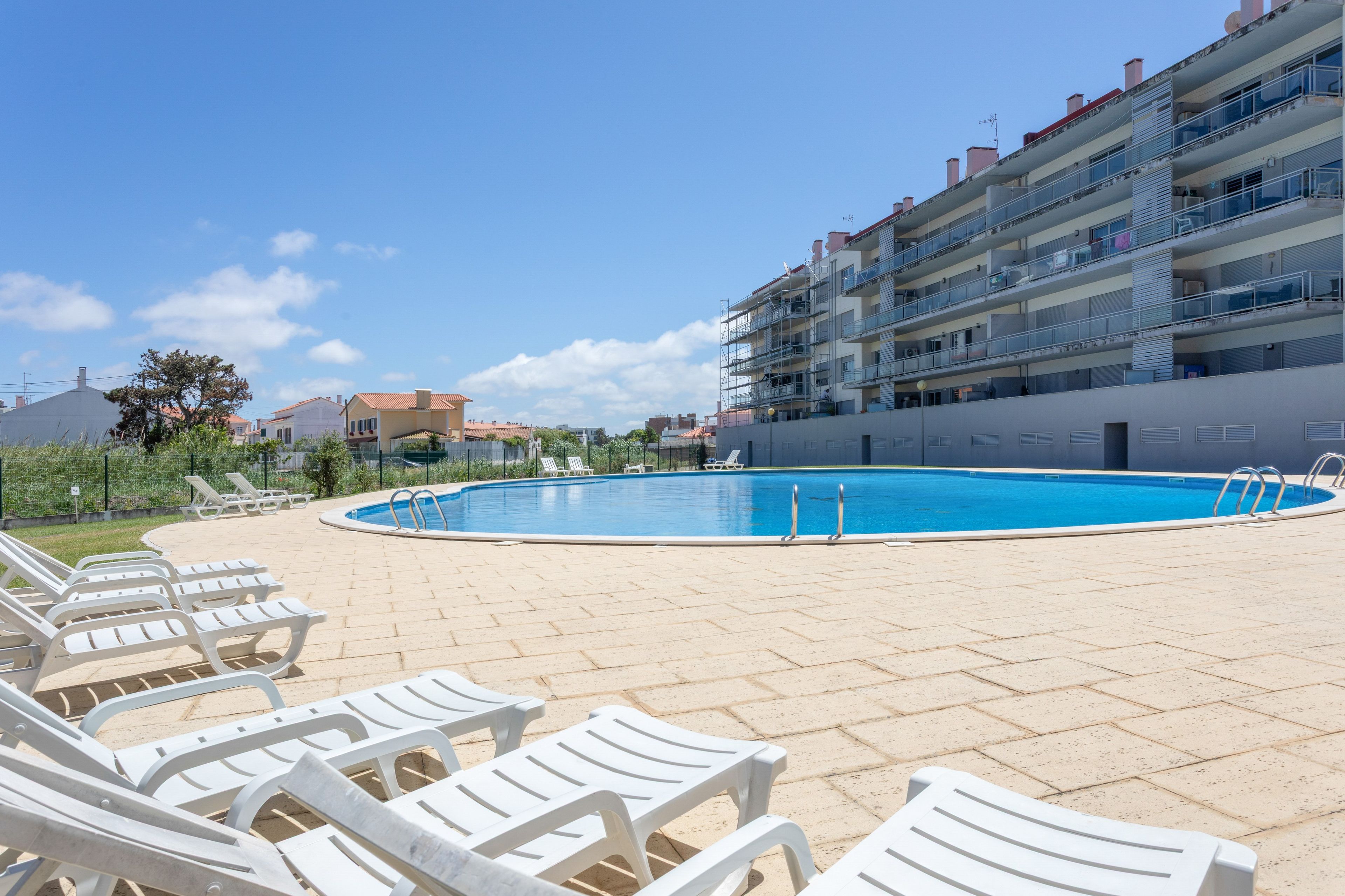 A swimming pool with lounge chairs, a clear sky, and a multi-story building in the background.