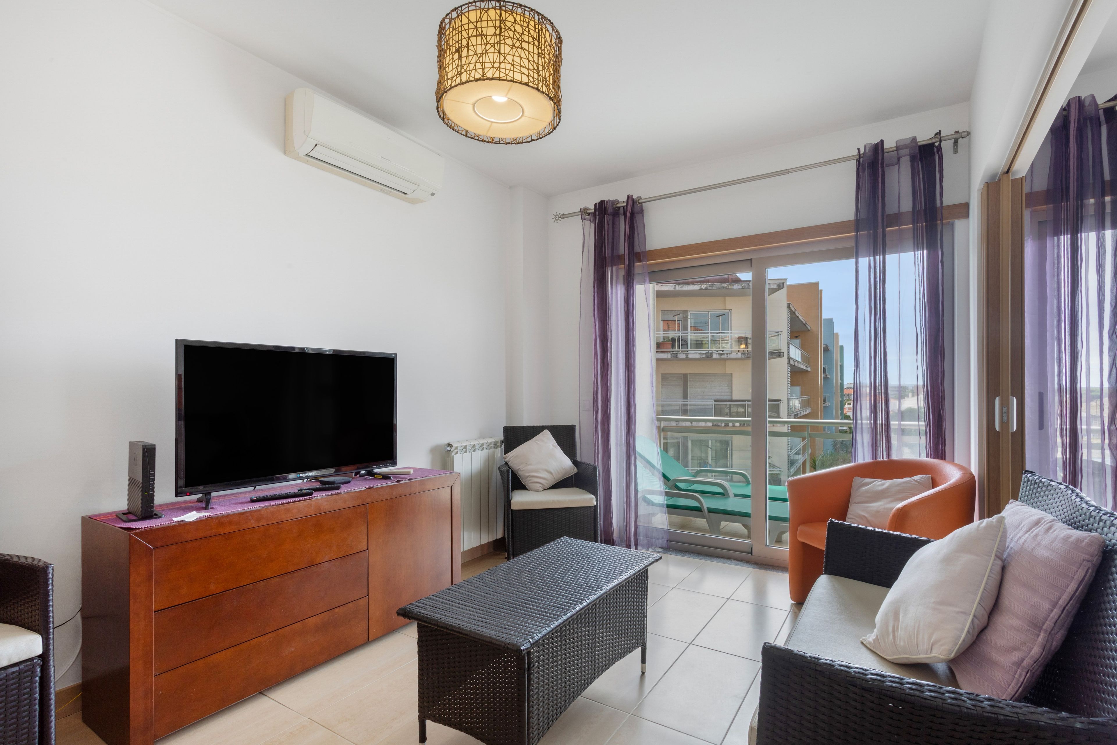 Ein modernes Wohnzimmer mit Fernseher, Holzschrank, Rattan-Sofa und einem Balkon mit lila Vorhängen.