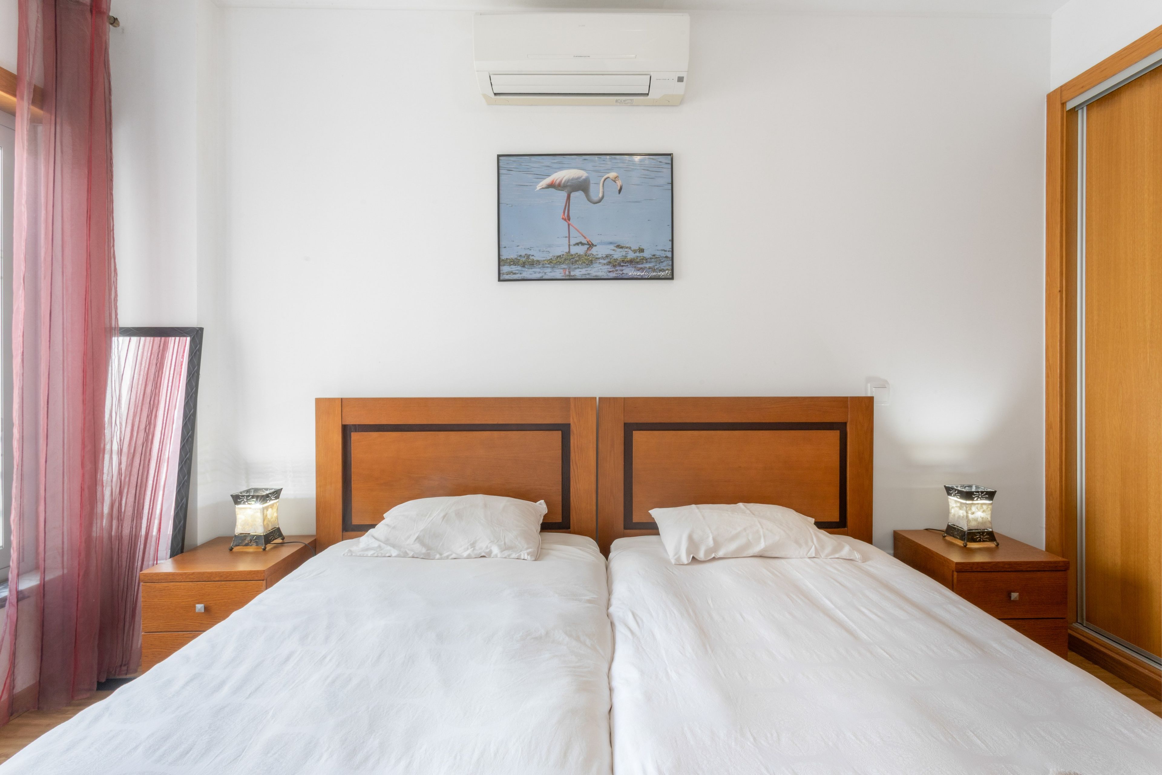 Una cama doble con cabecera de madera, dos mesitas de noche con lámparas, un aire acondicionado de pared y un cuadro con una imagen de un flamenco.
