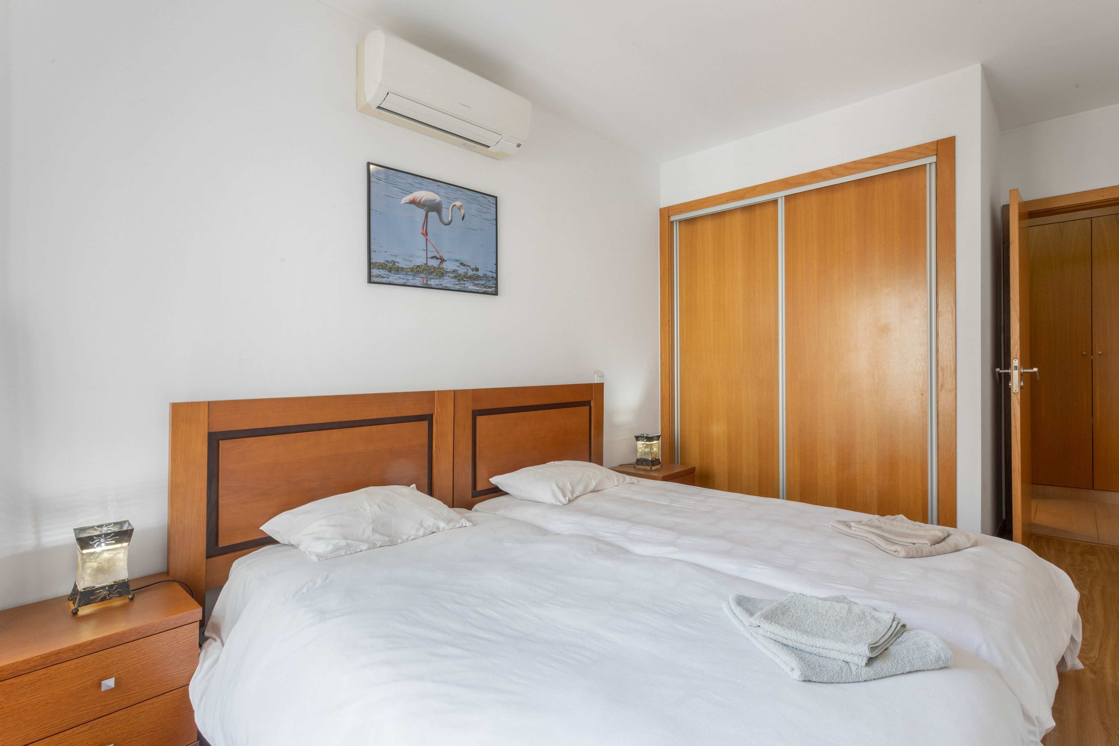 Une chambre d’hôtel avec une tête de lit en bois, un lit avec du linge de maison blanc, une table de chevet avec une lampe, une armoire en bois et un climatiseur fixé au mur.
