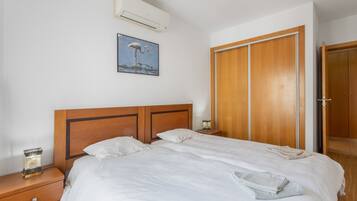 Une chambre d’hôtel avec une tête de lit en bois, un lit avec du linge de maison blanc, une table de chevet avec une lampe, une armoire en bois et un climatiseur fixé au mur.