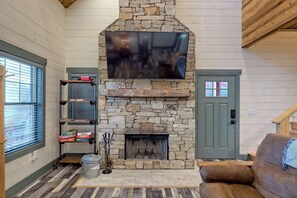 A living room with a stone fireplace, a mounted flat-screen TV, and shelves with board games.