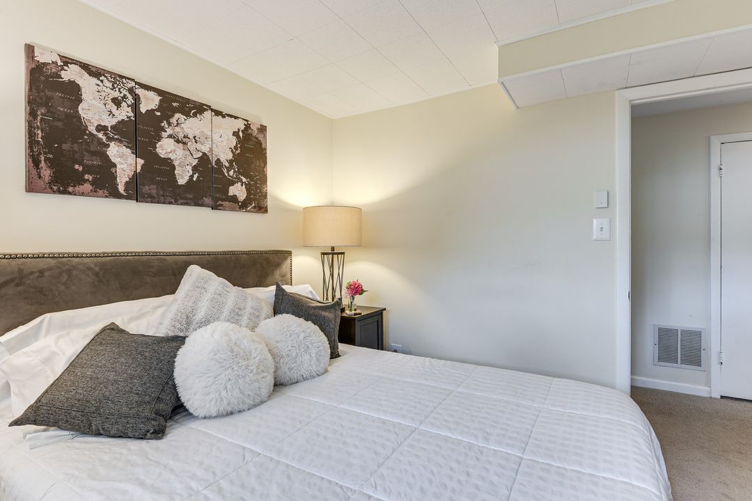 A neatly made bed with a white comforter and multiple pillows, a world map artwork on the wall, a bedside lamp, and a door to another room.