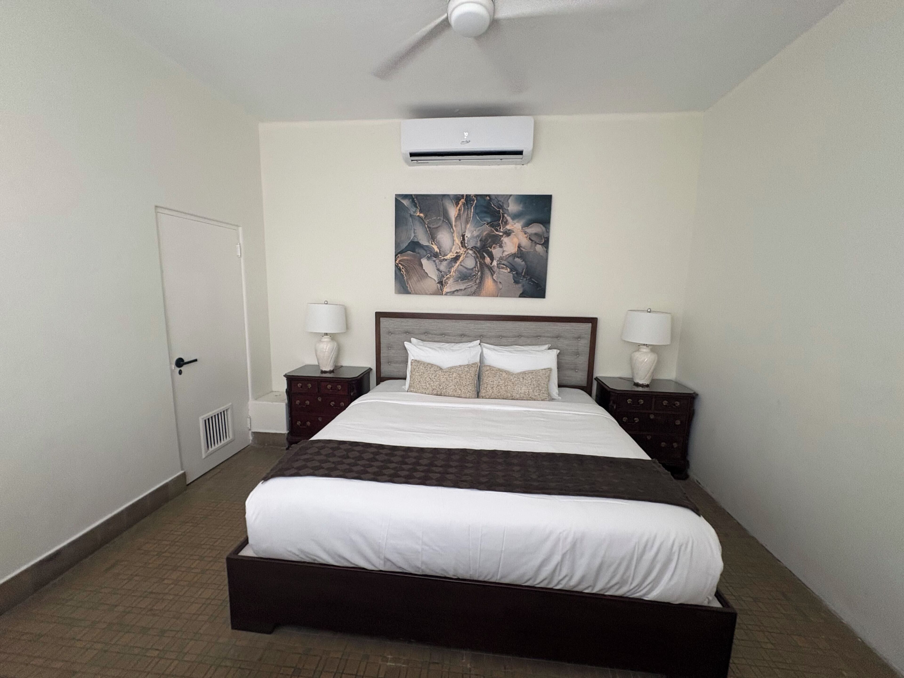 Una habitación de hotel con una cama, dos mesitas de noche, un ventilador de techo, un equipo de aire acondicionado y un cuadro en la pared.