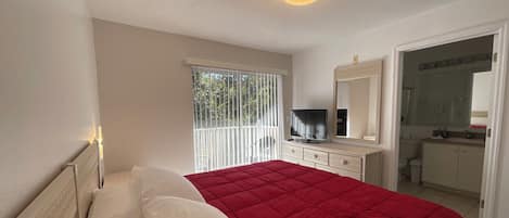 Une chambre à coucher avec un lit, une télévision, un miroir et une salle de bain visible à travers une porte ouverte.