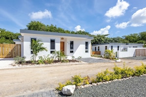 A modern two-story residential building with a white exterior, a front entrance with a wooden door, and a small garden area with various plants. - Prime Location: New Home in Jan Thiel with Private Pool (Willemstad)