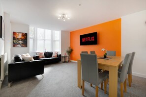 Ein modernes Wohnzimmer mit einer schwarzen Couch, einem Holzesstisch mit Stühlen und einer großen, orangefarbenen Wand mit einem Netflix-Schild.