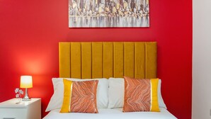Habitación de hotel con pared roja, un cuadro de un paisaje urbano, una cama con almohadas blancas y naranjas, y una mesita de noche con una lámpara.